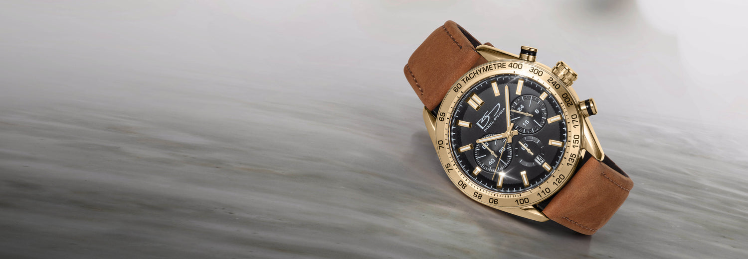 Close-up of a luxury gold chronograph watch with a black dial and brown leather strap resting on a textured gray surface, highlighting intricate details and craftsmanship.
