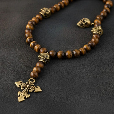 Necklace with brown beads and gold skull and cross pendant on a dark background. Daniel Steiger Skull Tiger Eye Rosary
