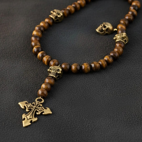 Necklace with brown beads and gold skull and cross pendant on a dark background. Daniel Steiger Skull Tiger Eye Rosary