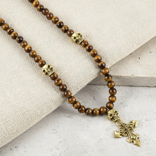 Beaded necklace with skull and cross design on a textured surface. Daniel Steiger Skull Tiger Eye Rosary