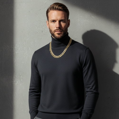 Man wearing a dark turtleneck sweater with a gold chain necklace against a gray wall.