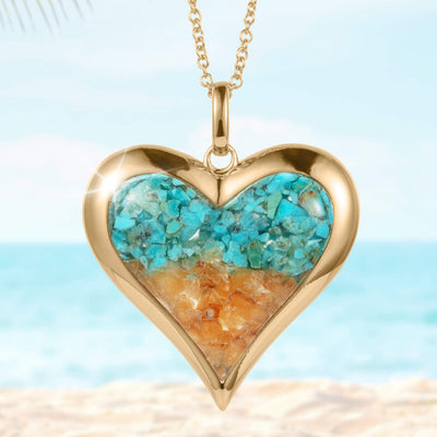 Heart-shaped gold pendant necklace from the Sunlit Shore Heart Collection featuring blue and sandy-colored stone inlay, displayed against a beach background.