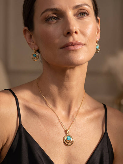 Woman wearing layered gold necklaces with a turquoise pendant and matching turquoise drop earrings against a warm indoor background